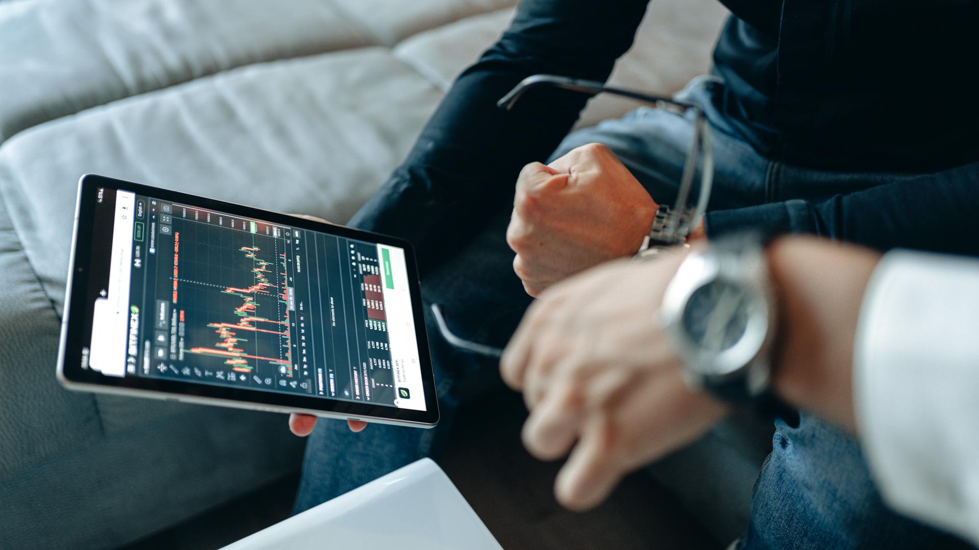 Close-up of tablet with trading charts, held by people analyzing online trades indoors.