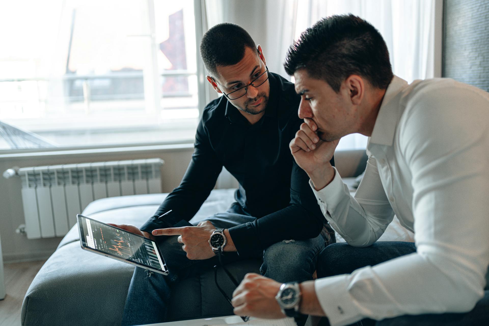 Two men collaborating in a home office, analyzing financial charts on a tablet.