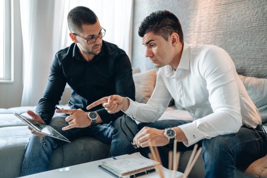 Two men in a home office discuss work on a tablet, highlighting teamwork and collaboration.