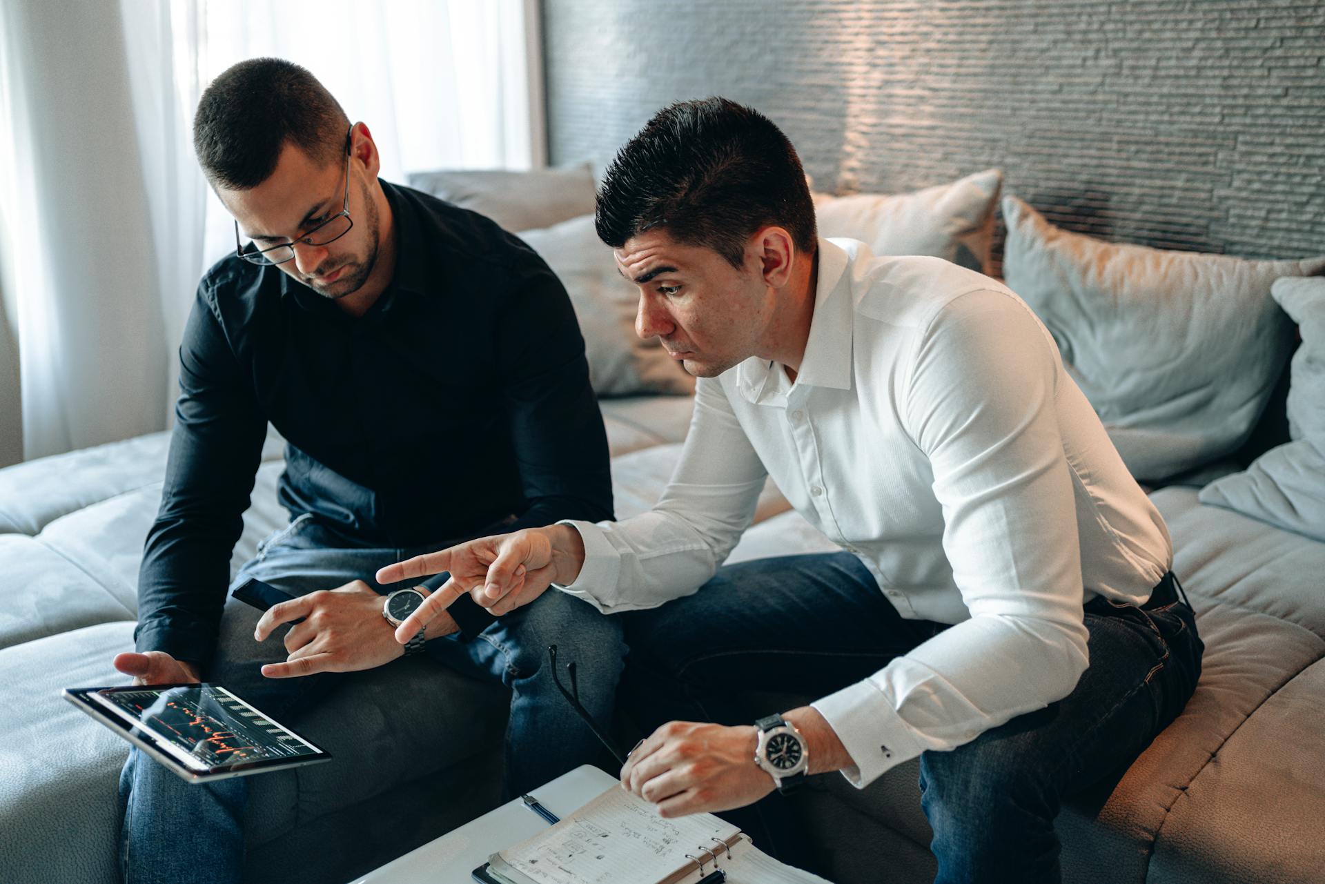 Two businessmen discussing financial data on a digital tablet while sitting on a couch.