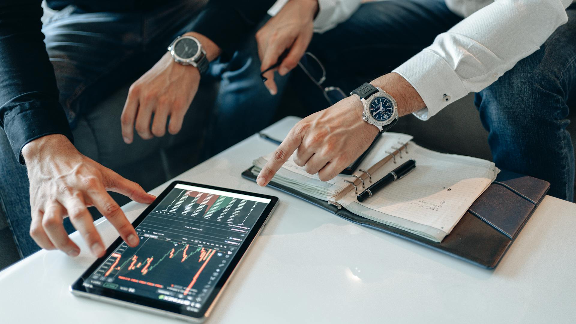 Close-up of professionals discussing stock charts on a tablet with notes and pens on a table.