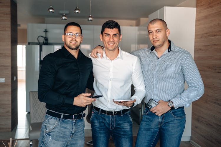 Three Men In Long Sleeve Shirt Standing Together Holding Gadgets