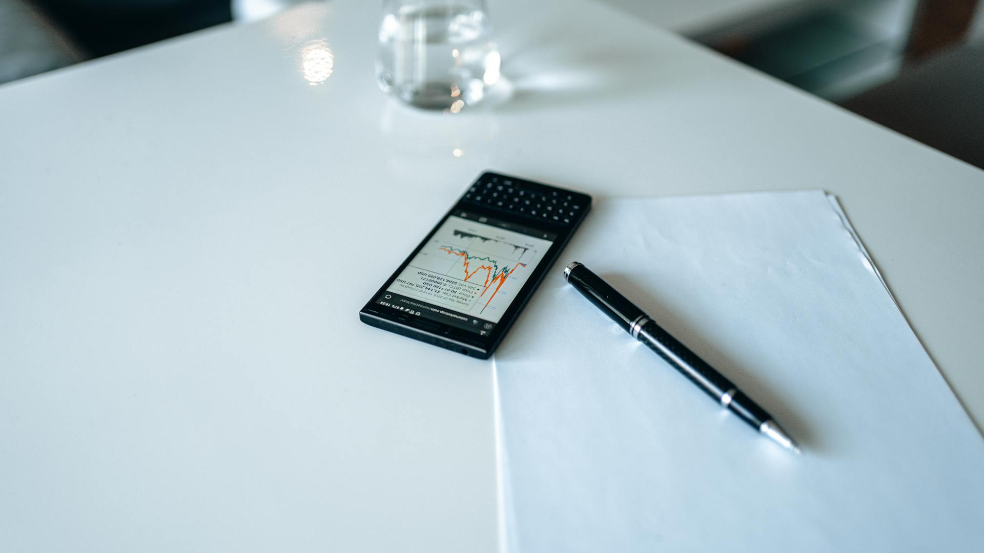 Smartphone displaying stock market graph on white desk with pen and paper.