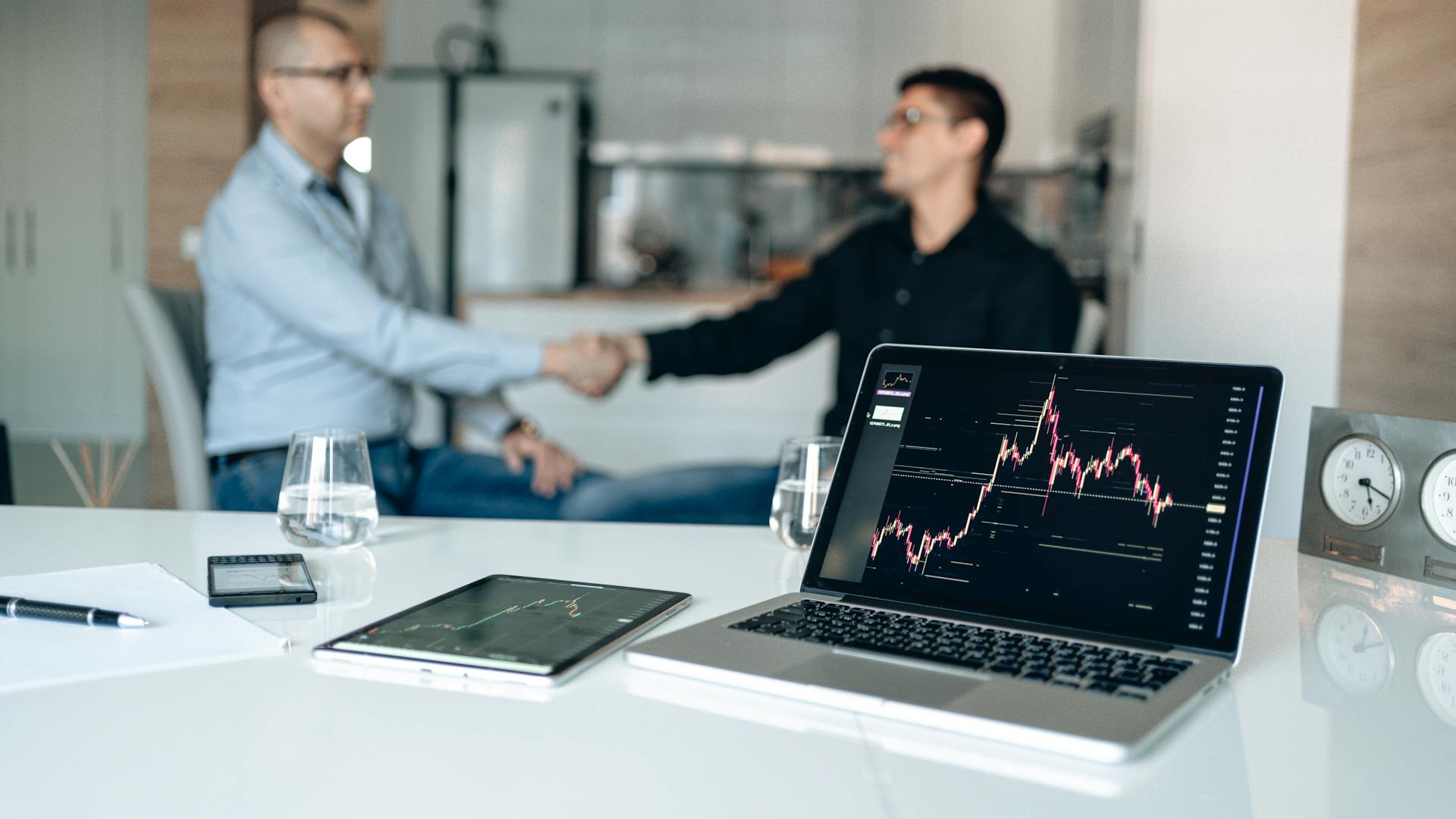 Two businessmen shake hands at a desk with stock market graphs on a laptop.