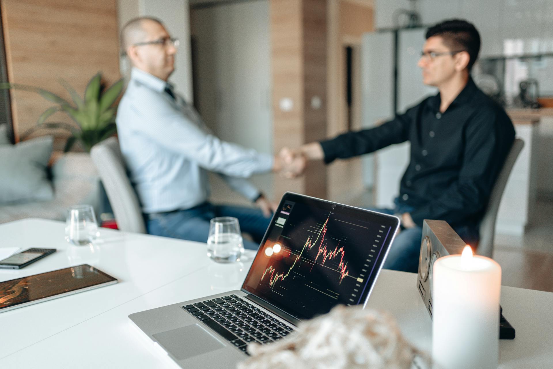 Two businessmen shaking hands in a modern office with trading data displayed on a laptop.