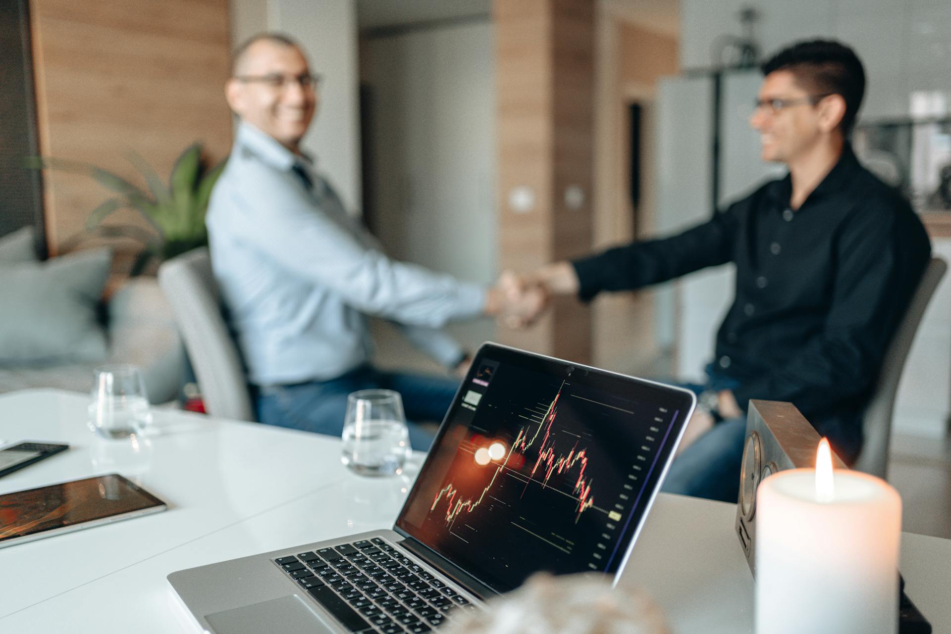 Two businessmen handshake over a table with a laptop showing financial graphs in an office setting.