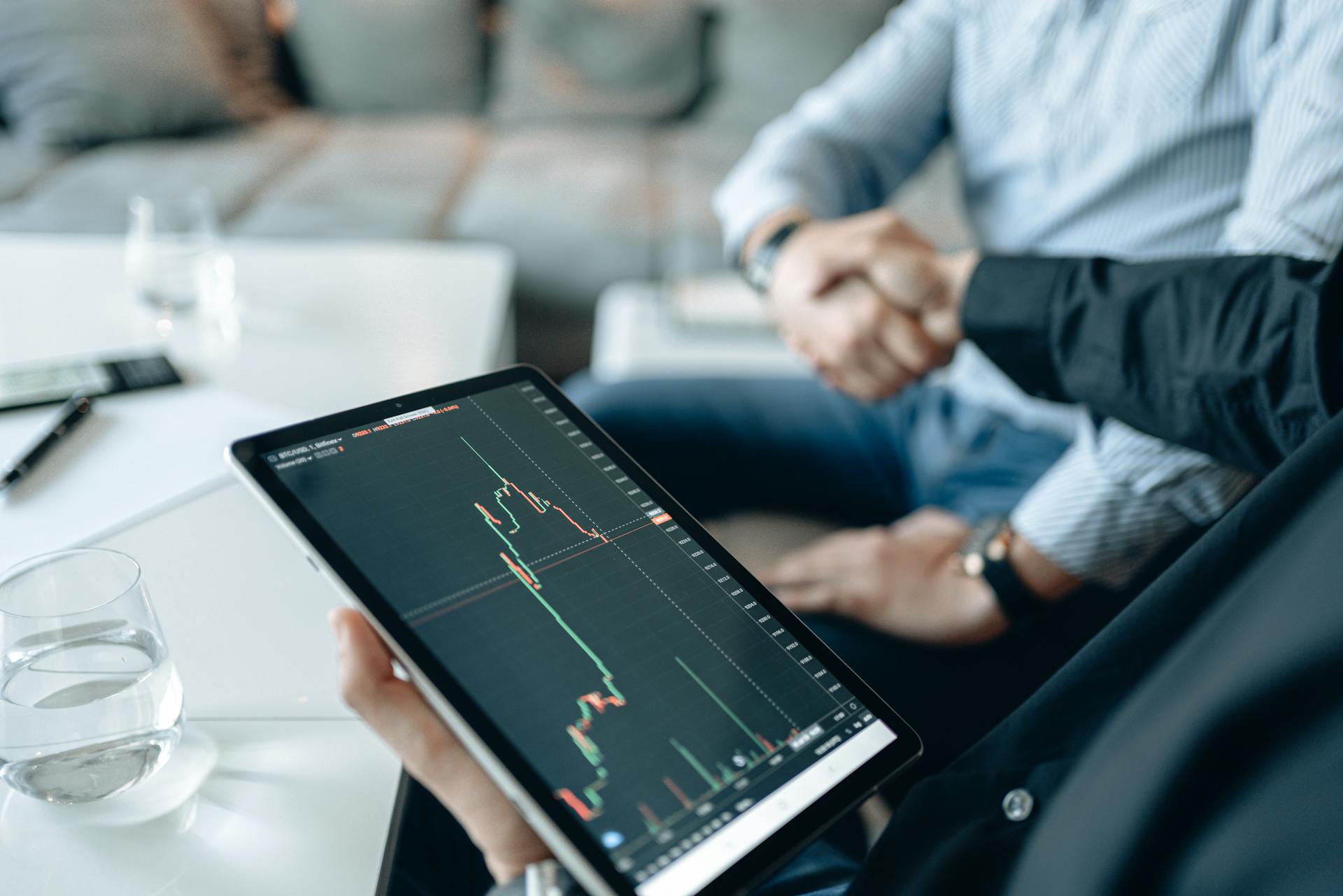 Close-up of tablet showing stock graph during business meeting with handshake in background.