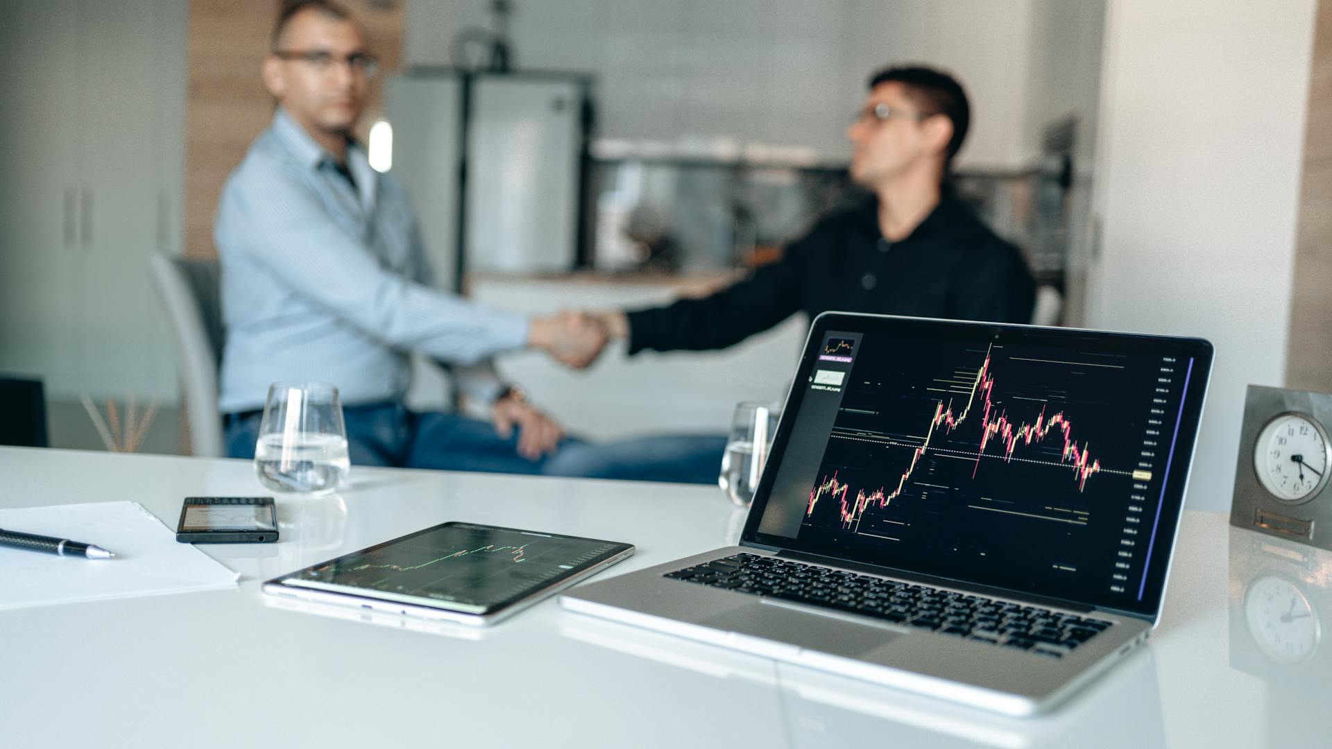 Two businessmen shaking hands in an office setting, with laptops displaying financial graphs.