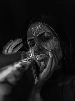 Black and white of serious female with neon paints on face looking at camera while standing near construction on black background
