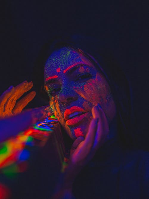 Calm female with glowing fluorescent paints on face looking at camera while standing on black background near construction in dark room