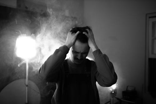Black and white of upset male standing in room with lightning equipment and smoke while thinking about difficult decision at home