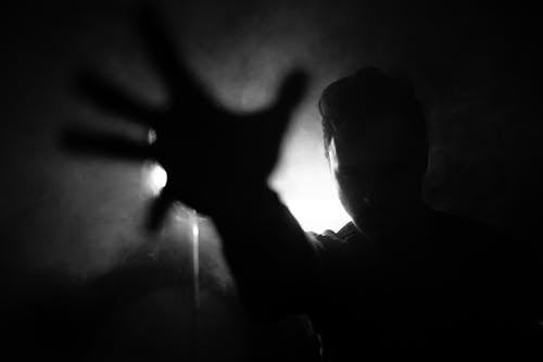 Black and white of anonymous male reaching out hand to camera while standing near wall with lamp in dark room