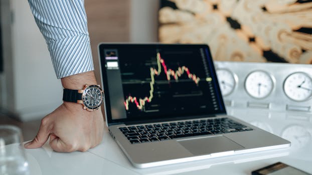 A sophisticated workspace featuring a laptop with a stock chart and a watch on a man's wrist.