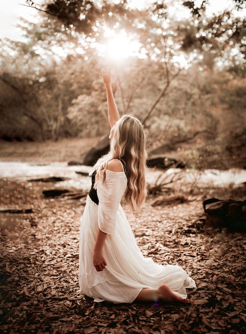 Free A woman kneels in a forest, reaching towards the sun, with fallen leaves and serene ambiance. Stock Photo