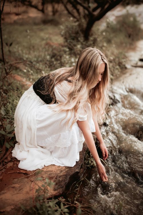 Free A woman in a white dress gently touching a stream surrounded by nature. Stock Photo