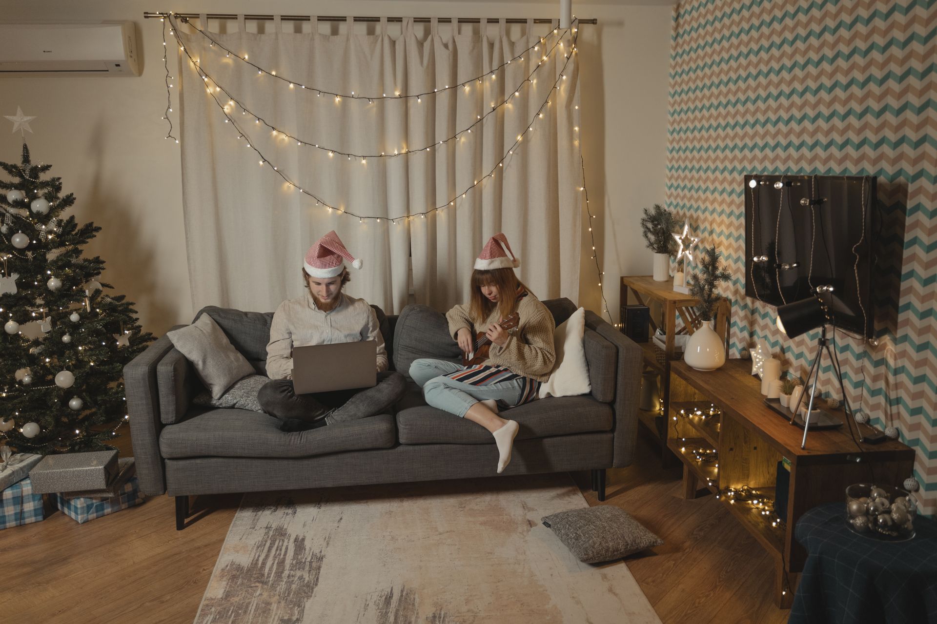 A festive living room scene featuring a couple wearing Santa hats, using a laptop and playing a ukulele.