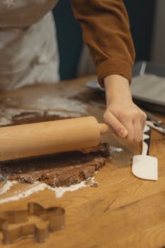 A person rolls brown dough on a wooden table with a rolling pin, preparing for baking.