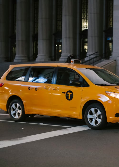 Modern shiny yellow minivan taxi driving on asphalt road near building with columns in daytime