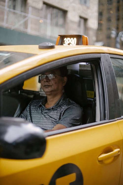 Serious middle aged ethnic man in polo shirt and eyeglasses driving modern yellow taxi car on city street in daytime