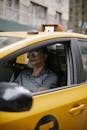 Serious middle aged ethnic man in polo shirt and eyeglasses driving modern yellow taxi car on city street in daytime