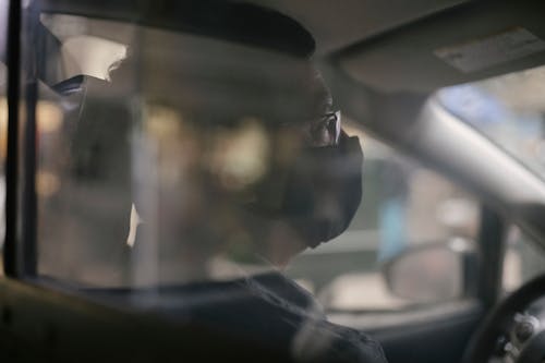 Free Through glass of crop concentrated ethnic man in eyeglasses sitting in car and wearing medical mask because of coronavirus pandemic Stock Photo