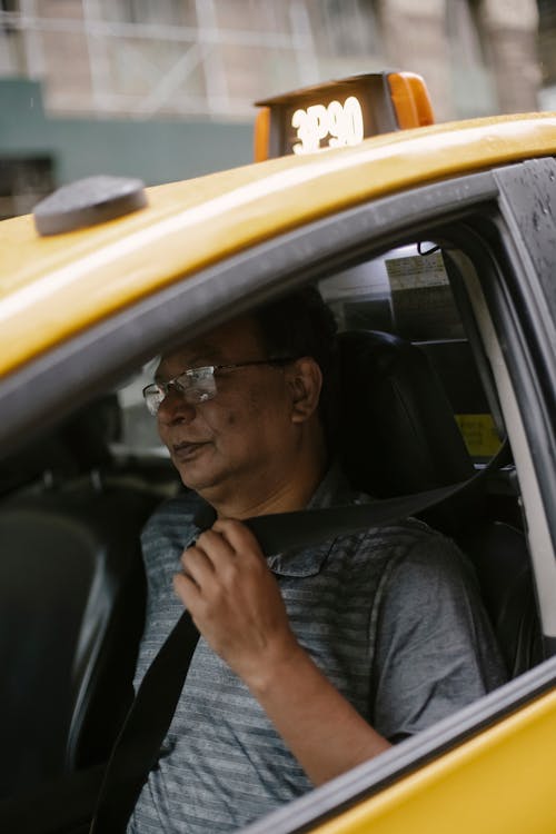 Free Serious ethnic mature man in casual clothing fastening safety seatbelt before driving modern yellow taxi car on blurred background Stock Photo