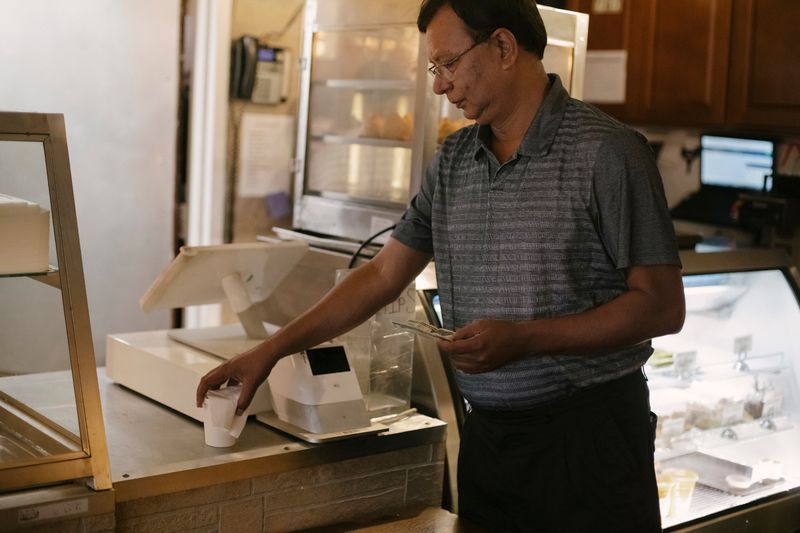 Side view of Asian male wearing eyeglasses standing near cash register and showcase while piking up cup of coffee in cafe