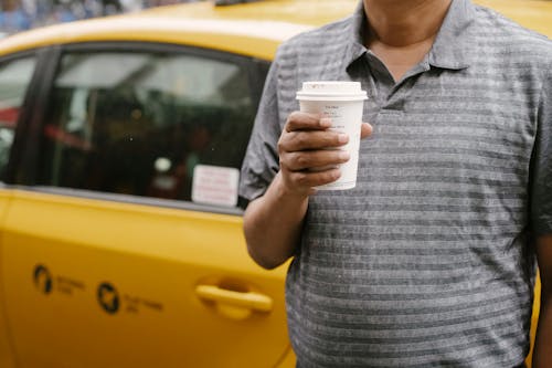 Unrecognizable male in casual outfit with cup of takeaway coffee standing near yellow taxi on city in street during break