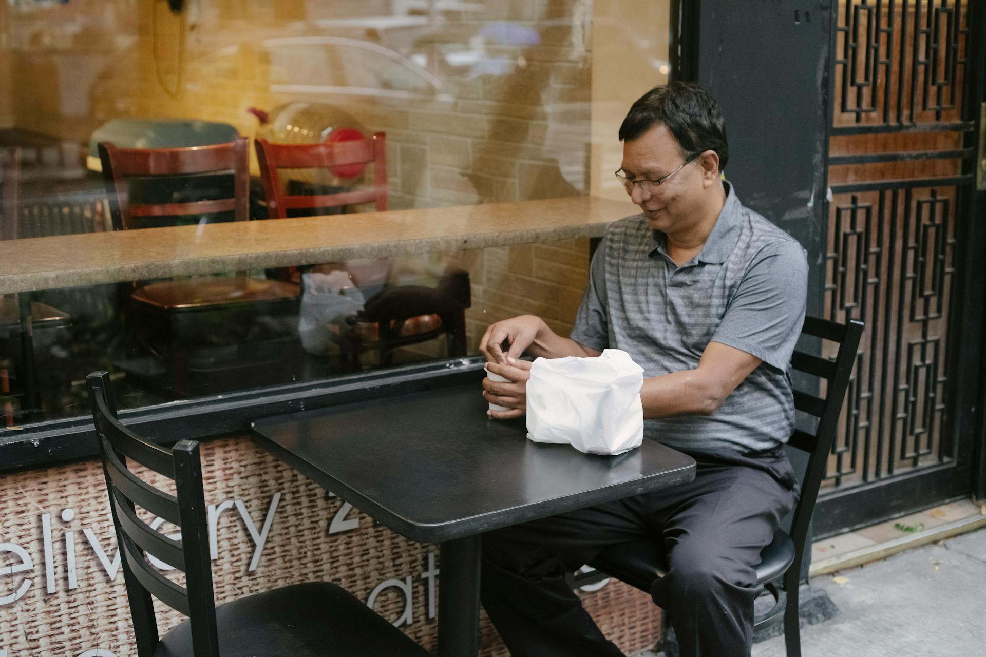 Cheerful Asian male sitting near window on street cafeteria at table with plastic bag and paper cup of coffee in hands