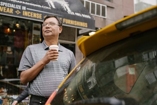 一位戴眼镜、神情严肃的亚裔中年男子，拿着外卖热饮站在街上，目光移开，身旁是黄色的出租车和咖啡馆