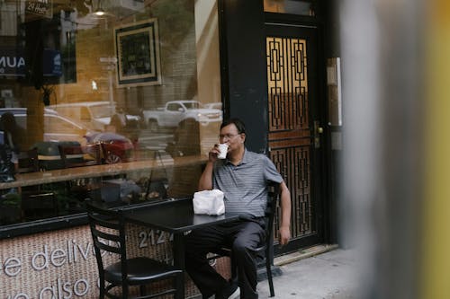 Serious ethnic male in eyeglasses sitting at table near window in street cafe while drinking hot beverage from cup during coffee break