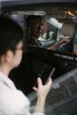 View through open window of ethnic taxi driver looking over shoulder on passenger in glasses surfing smartphone