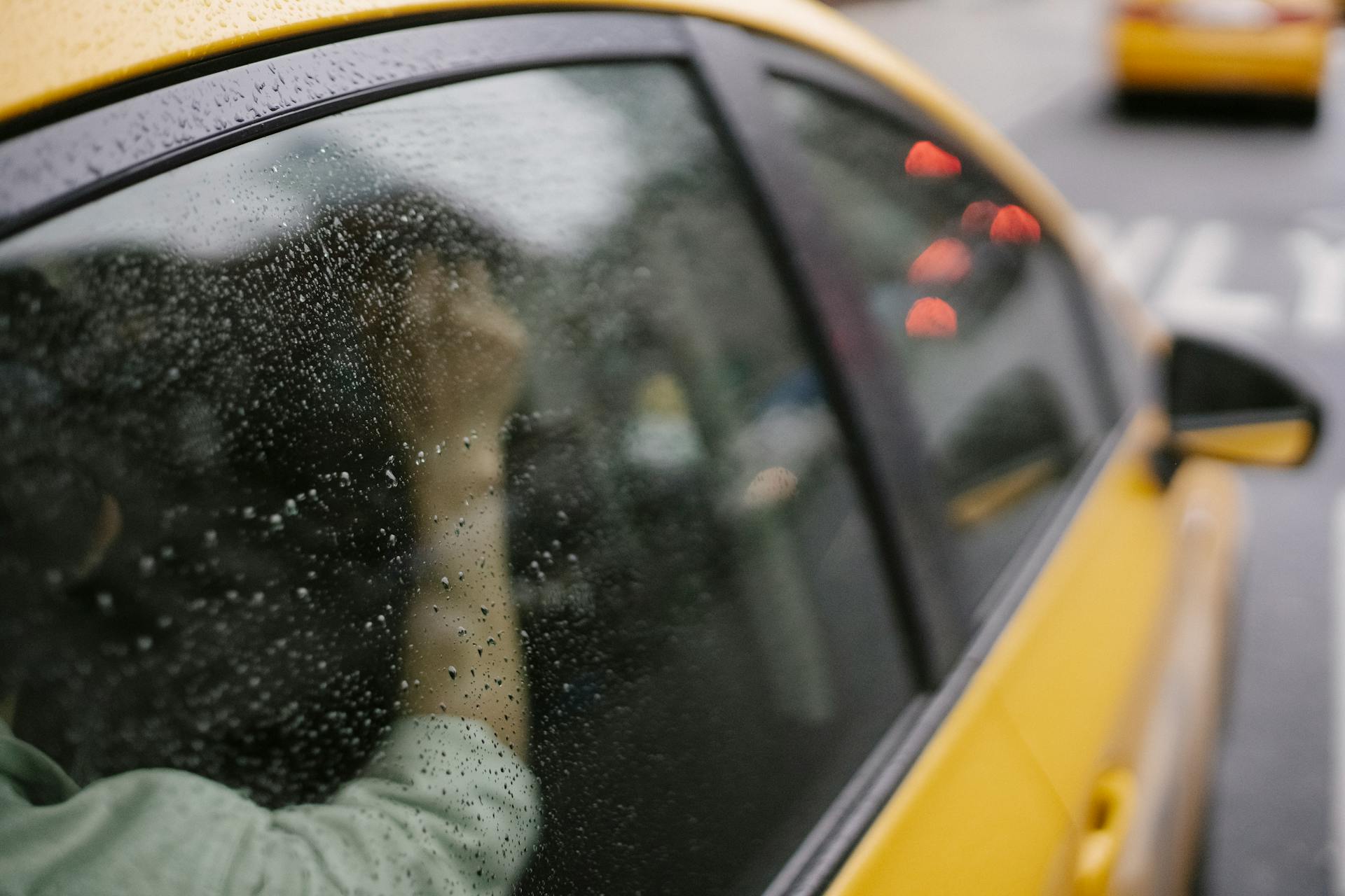 Crop anonymous passenger sitting in yellow taxi car on road in town in rainy weather