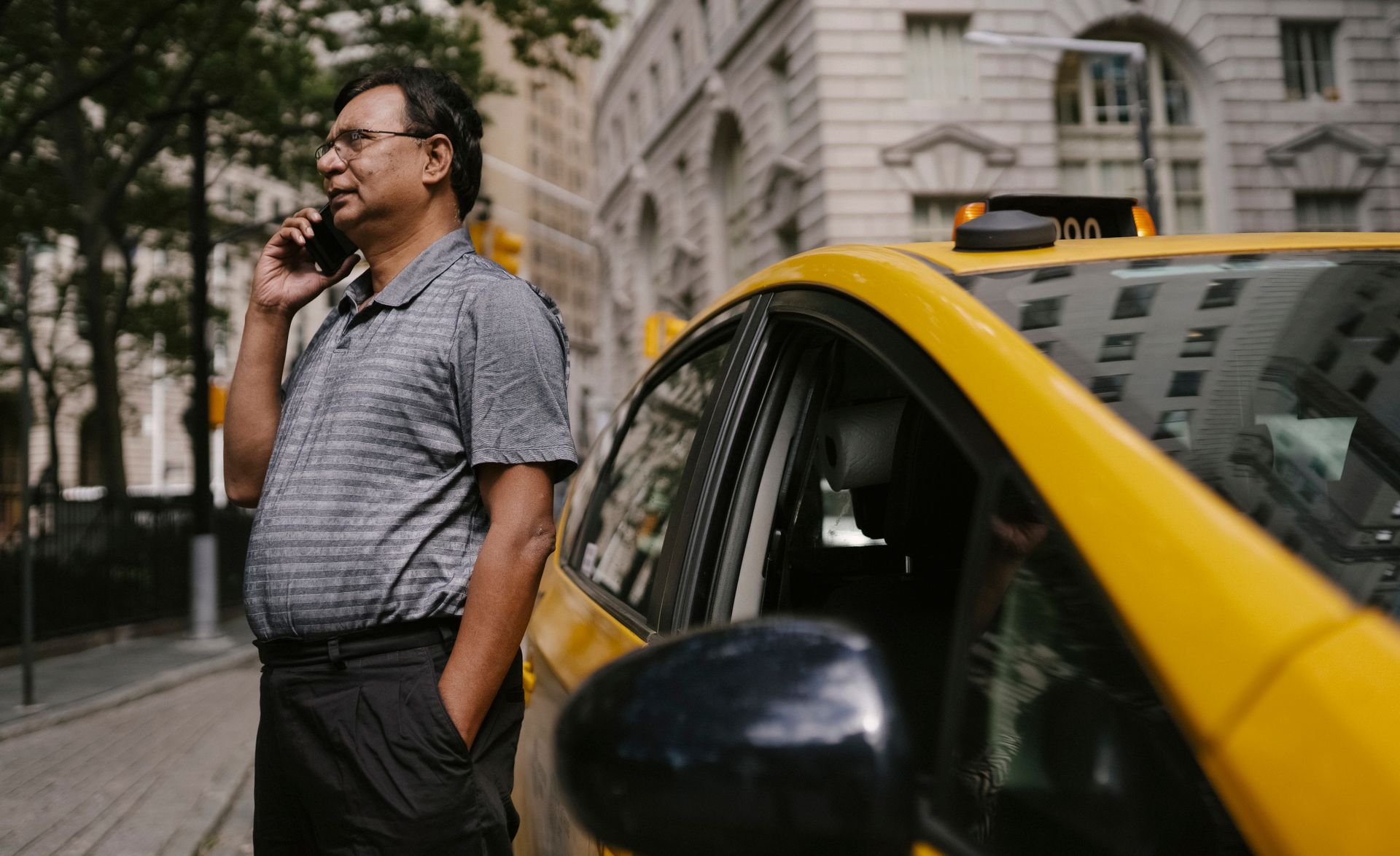 Serious ethnic male taxi driver with hand in pocket having conversation on cellphone while standing near car