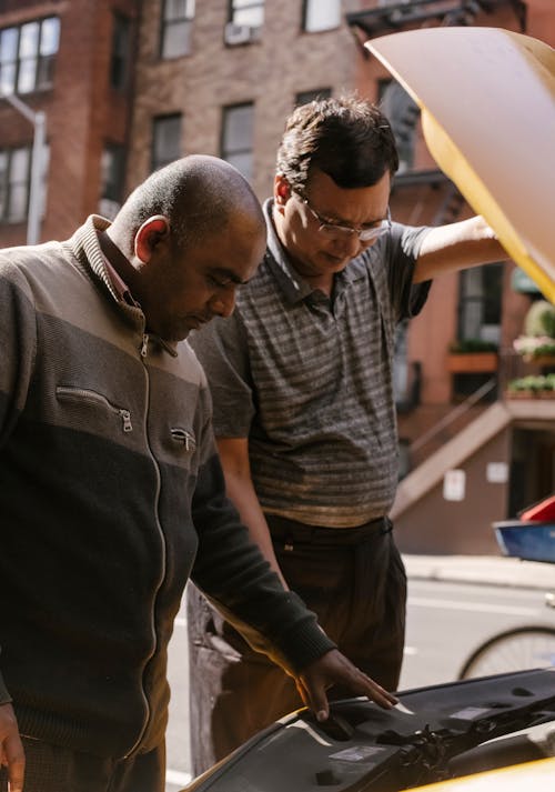 Serious ethnic man looking at motor and details of car with opened hood parked on roadside