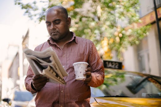 Interested adult ethnic male taxi driver with disposable glass of hot drink reading newspaper near vehicle on city street