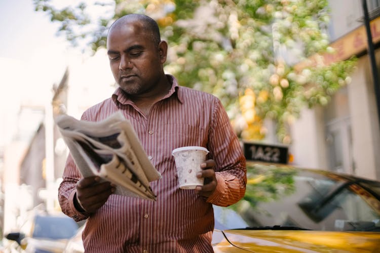 Serious Ethnic Cab Driver With Takeaway Coffee And Newspaper