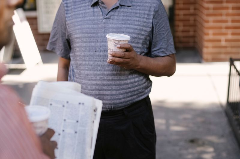 Crop anonymous ethnic male friends with coffee to go and newspaper speaking on city pavement