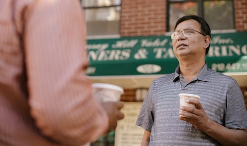 Attentive middle aged Asian male in eyeglasses with takeaway coffee talking to crop anonymous partner on city street