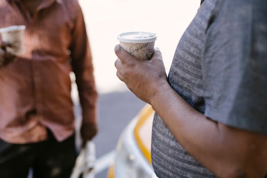 Crop unrecognizable ethnic male coworkers with takeaway coffee conversing near taxi car on urban roadway