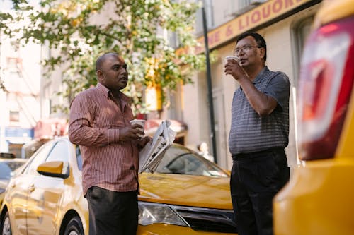 Free Multiethnic partners with coffee to go and newspaper conversing near taxi cars during break from work in town Stock Photo