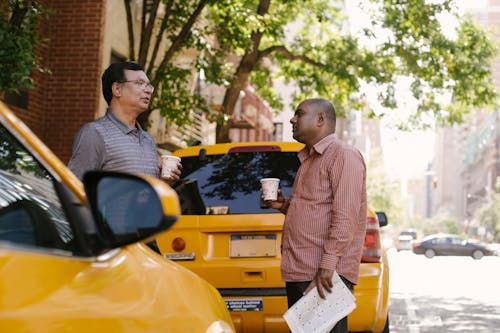 Free Side view of multiracial male coworkers with newspaper and takeaway coffee speaking near taxi autos while looking at each other in town Stock Photo