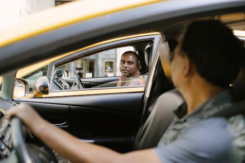 Free Side view of adult ethnic male cab driver interacting with anonymous colleague driving auto while looking at each other in city Stock Photo