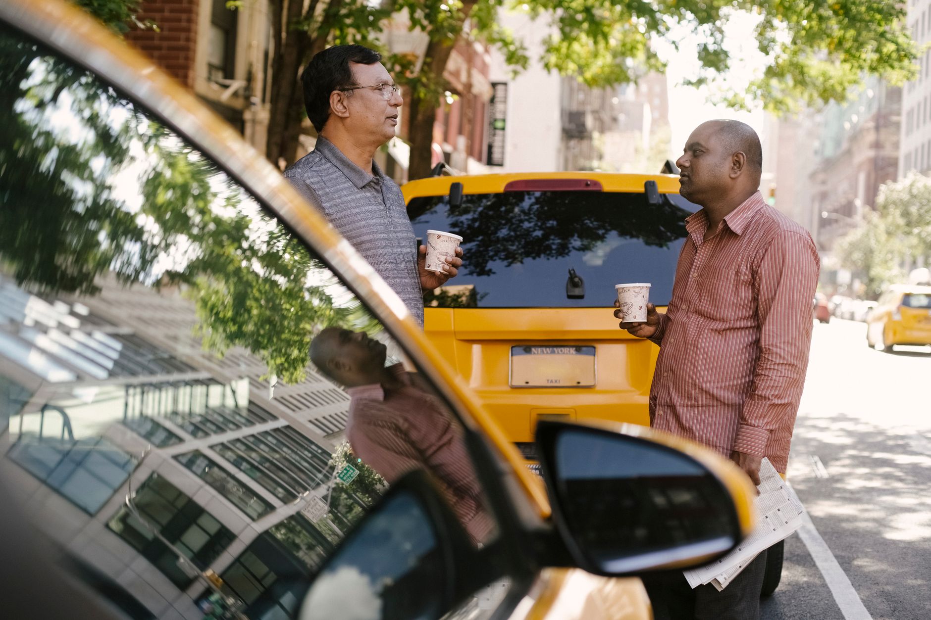 Side view of multiracial male coworkers with takeaway coffee conversing near yellow automobiles on urban road while looking at each other