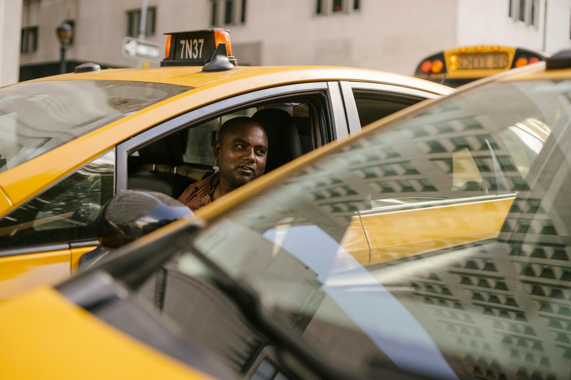 Adult ethnic male taxi driver sitting in contemporary auto and looking away near similar vehicle reflecting urban building