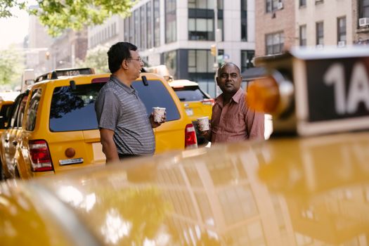 一名多种族男性出租车司机端着一次性热饮，在城市的黄色汽车旁讲话