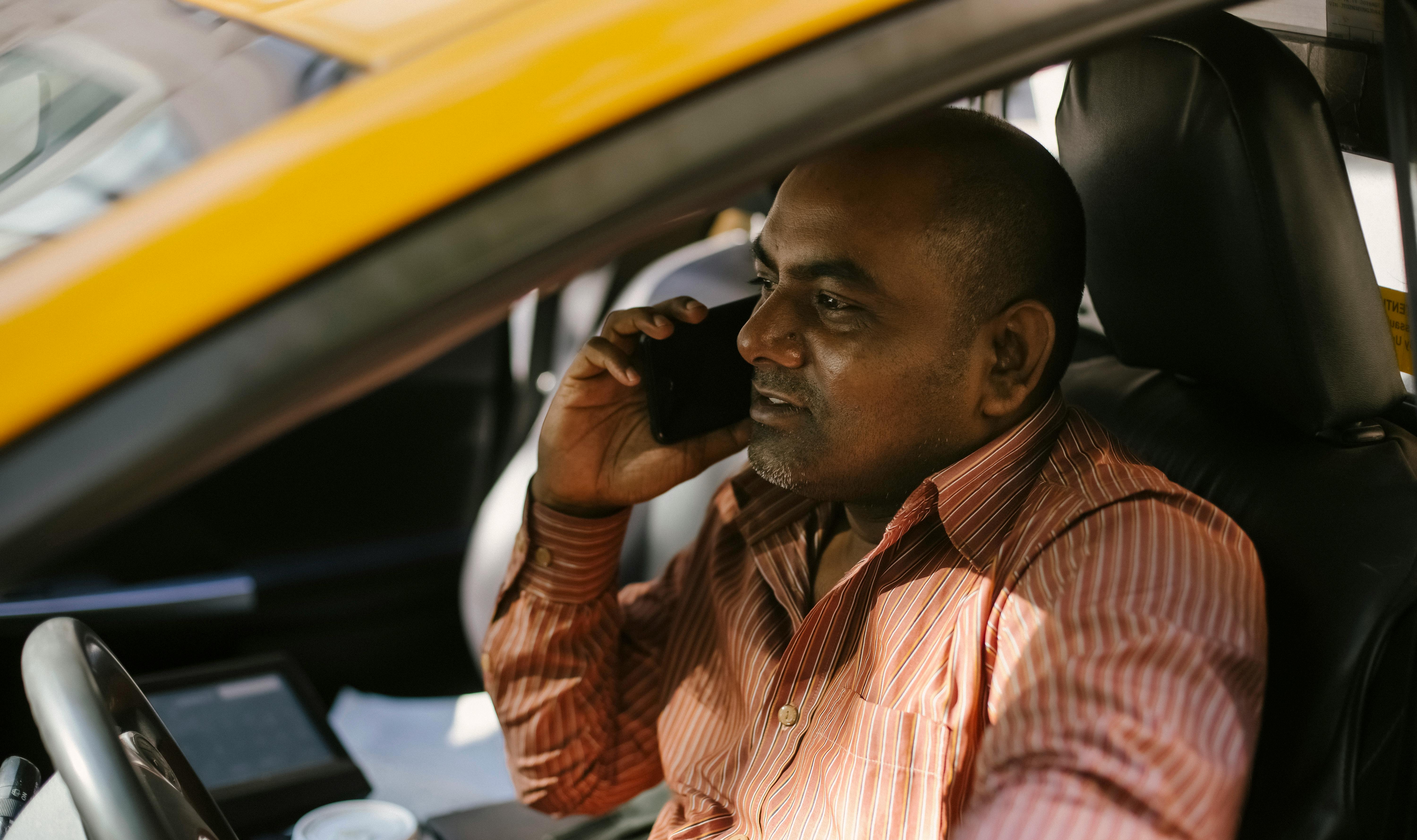 Ethnic male cab driver in striped shirt talking on cellphone in transport while looking away