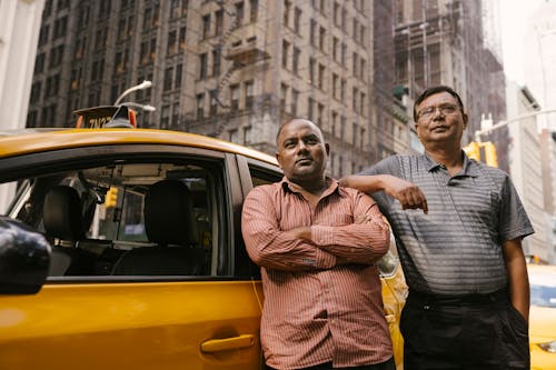 Ethnic taxi driver with crossed arms near mature Asian colleague with hand in pocket looking away near automobile in city