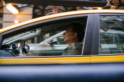 Free Side view of ethnic male driver sitting behind steering wheel driving yellow cab with lowered window on city in street with blurred background Stock Photo