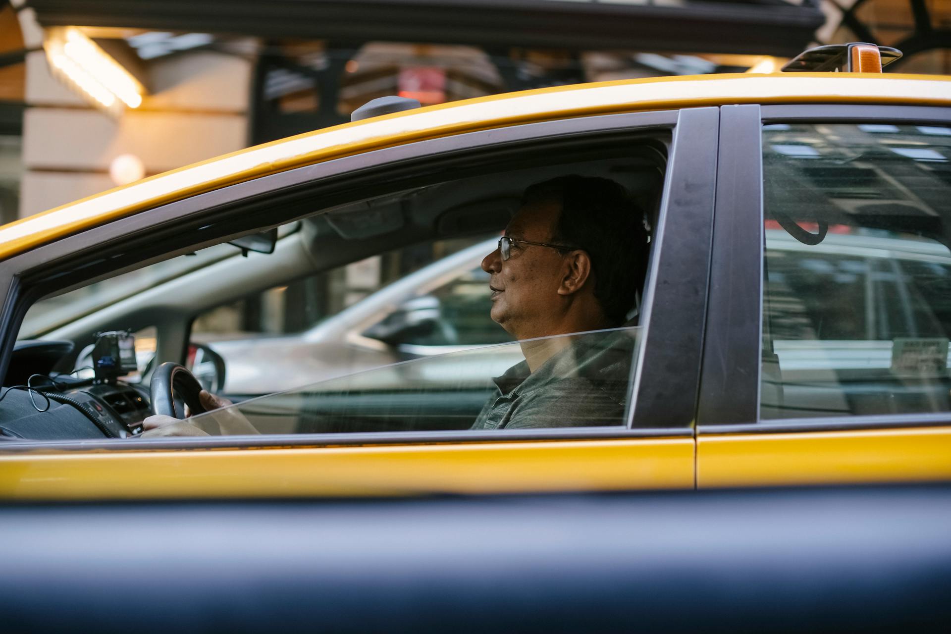 Side view of ethnic male driver sitting behind steering wheel driving yellow cab with lowered window on city in street with blurred background